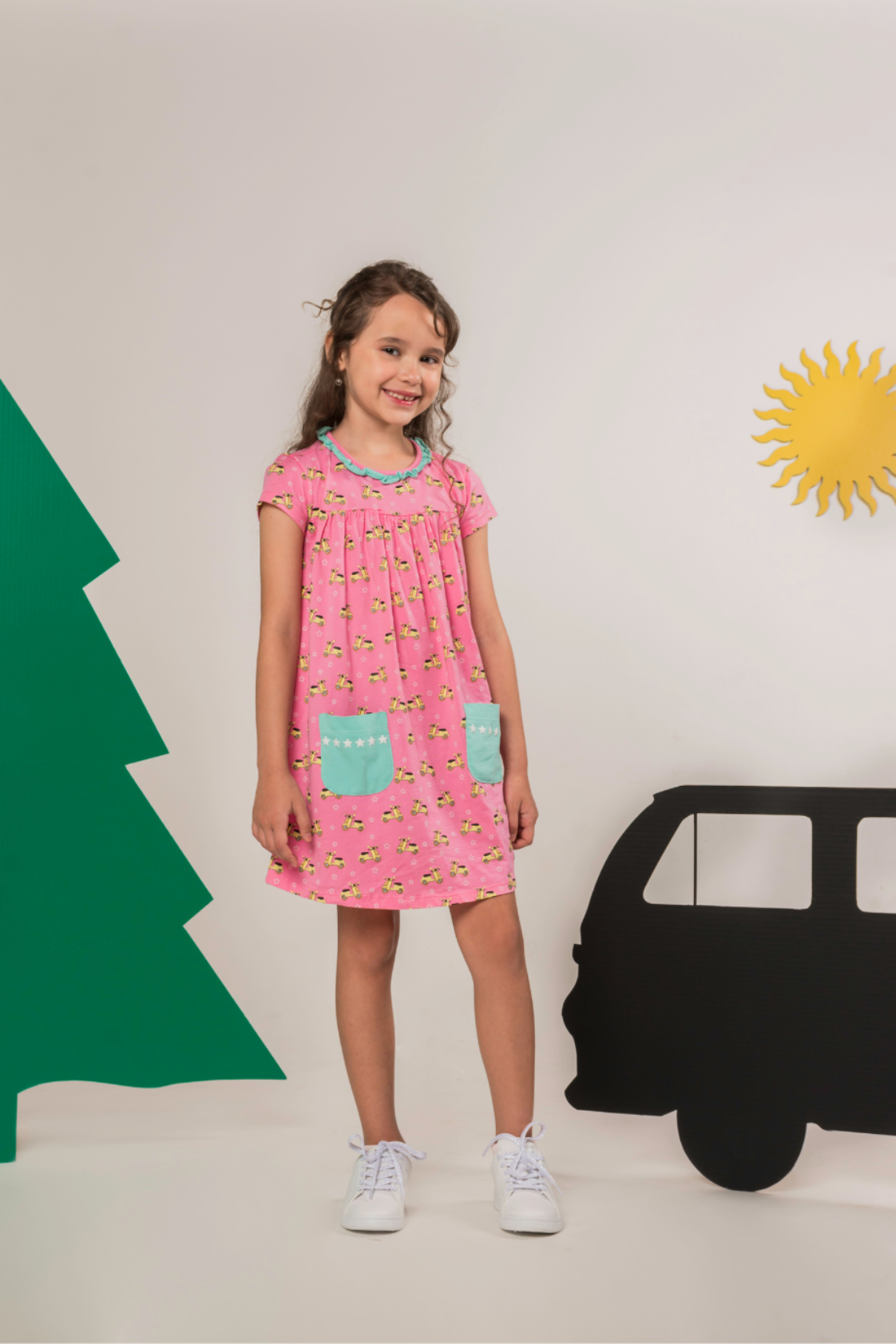 Young girl smiling in a pink empire-silhouette dress covered in scooter and star prints, featuring front pockets and a delicate ruffled neckline.