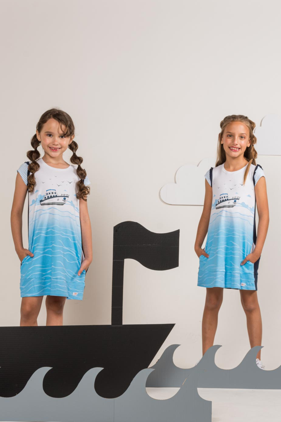 Two young girls wearing a white front, ocean-blue back dress with an illustrated sailboat print, featuring textured ruffle sleeves and side pockets.