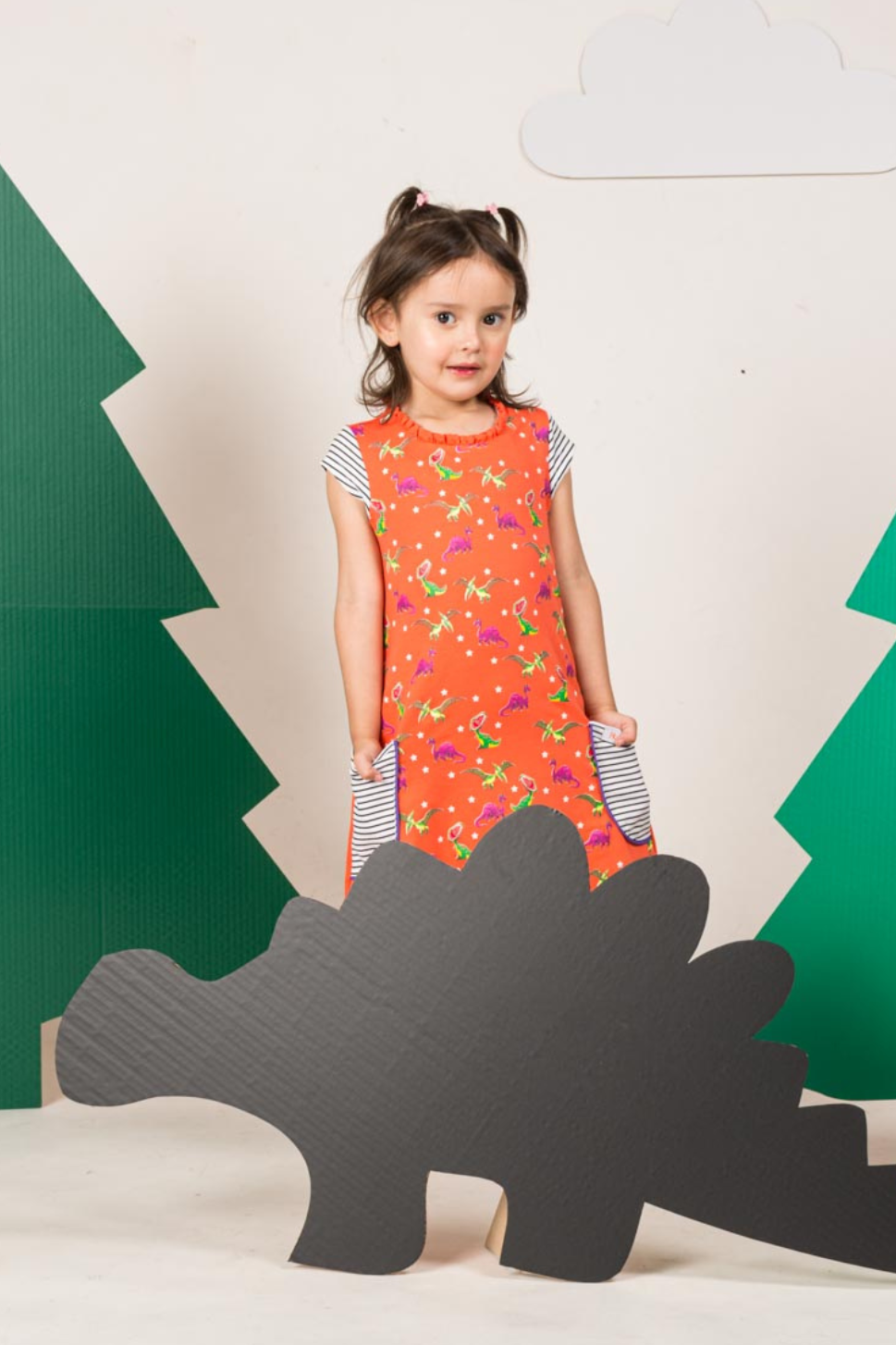Little girl exploring outdoors in a coral pink short-sleeve dress featuring a fade-resistant T-Rex print and horizontal striped front pockets.