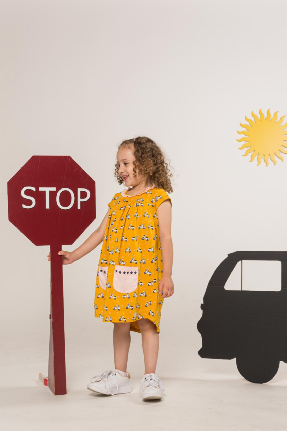 Cheerful yellow empire-silhouette dress for kids featuring a retro scooter print pattern, short sleeves, a ruffled collar, and star-lined pockets.